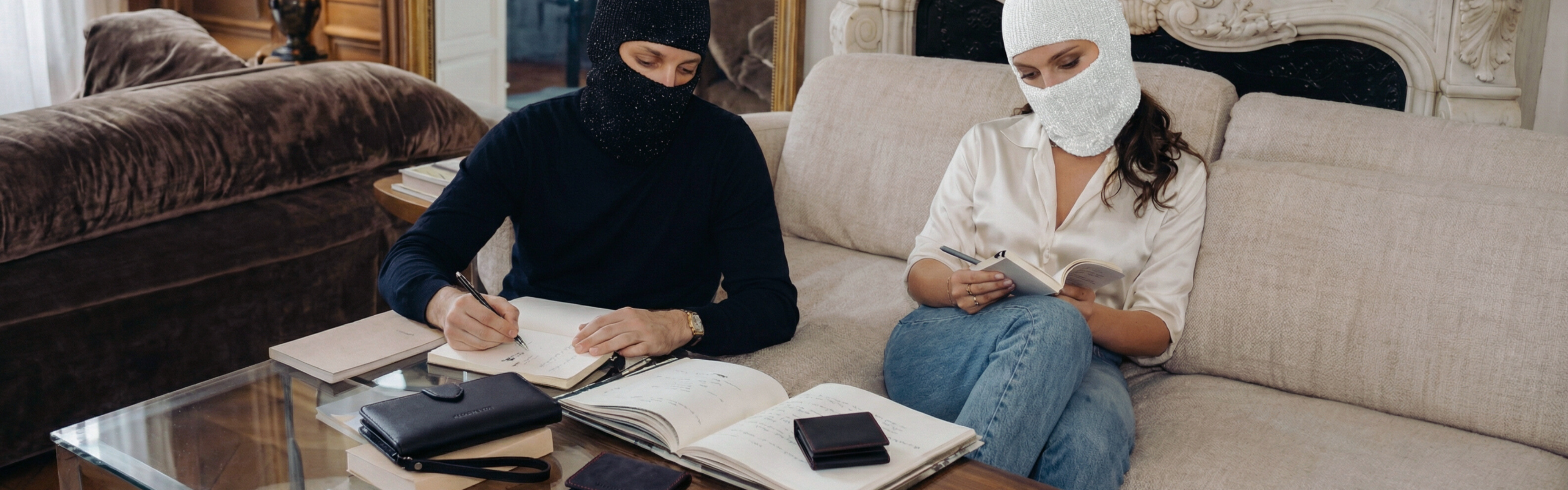 Masked man and woman sitting on a couch in a living room, one writing in a notebook and the other using a smartphone.